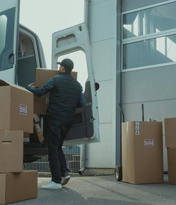 A delivery man unloading cardboard boxes from a van at a warehouse during the day.